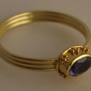 May include: A gold ring with a purple gemstone set in a bezel setting. The ring has a textured band with three grooves.