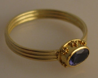 May include: A gold ring with a purple gemstone set in a bezel setting. The ring has a textured band with three grooves.