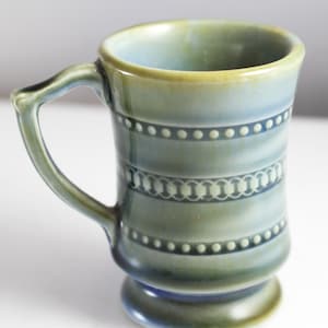 Puede incluir: Una taza de cerámica verde y azul con un asa blanca y un diseño celta. La taza tiene una base ancha y una parte superior estrecha.
