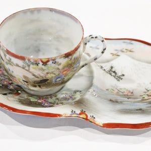 May include: A white porcelain teacup and saucer set with a floral design and a mountain scene. The saucer has a scalloped edge and a red rim.