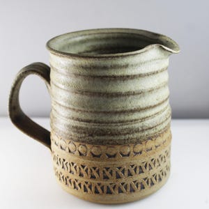 May include: A brown and green ceramic jug with a textured surface and a decorative pattern around the bottom. The jug has a wide mouth and a handle.