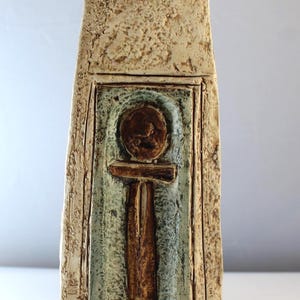 May include: A rectangular ceramic vase with a textured, off-white exterior. The center features a recessed panel with a brown ankh symbol against a blue-green background. The vase has a rustic, handcrafted appearance.