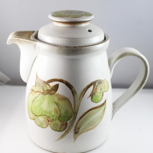 May include: A white ceramic coffee pot with a floral design in shades of green and brown. The pot has a lid with a small hole in the top.