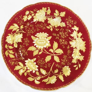 Op de afbeelding: Een rood dinerbord met een gouden bloemenontwerp. Het bord heeft een gegolfde rand en een gouden rand.
