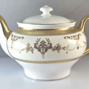 Könnte beinhalten: Weiße Porzellan-Teekanne mit goldenen Akzenten. Die Teekanne hat einen goldenen Griff, Ausguss und Rand. Ein goldenes Band mit floralen und Scroll-Designs umrundet den Körper. Der Deckel hat einen kleinen, weißen Knauf. Ein klassisches Stück für den Teeservice.