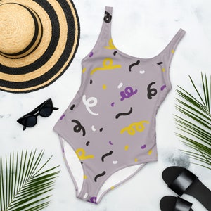 May include: A one-piece swimsuit with a light purple background and a pattern of yellow, white, black, and purple abstract shapes. The swimsuit is next to a straw hat, sunglasses, and sandals.