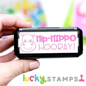 May include: A black and white self-inking stamp with a pink hippo illustration and the text "Hip-Hippo Hooray!"