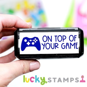 May include: A black and white self-inking stamp with a blue video game controller graphic and the text "ON TOP OF YOUR GAME".