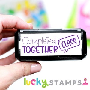 May include: A black self-inking stamp with white text that reads "Completed Together in Class" in a purple speech bubble. The stamp is held in a hand.
