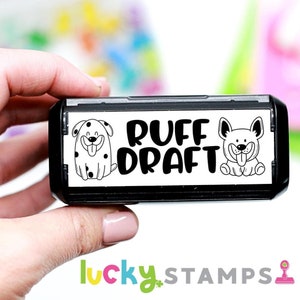 May include: Black and white self-inking stamp with a white background. The stamp features two cartoon dogs and the text "RUFF DRAFT".