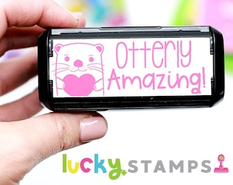 May include: A black and white self-inking stamp with a pink otter holding a pink heart and the text "Otterly Amazing!" in pink lettering.