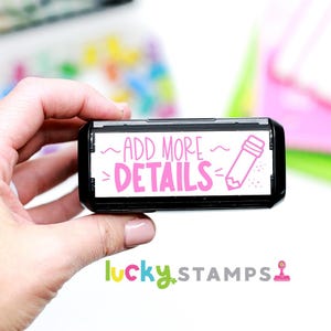 May include: A black and white self-inking stamp with pink text that reads "~ADD MORE~ DETAILS~" and a pink pencil icon. The stamp is held in a hand.