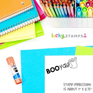 Puede incluir: Un sello de goma negro y blanco con un fantasma y el texto "BOOyah!" La impresi&oacute;n del sello es de aproximadamente 2,5 cm por 7 cm. El sello est&aacute; sobre una tarjeta blanca con un fondo azul y verde. La imagen tambi&eacute;n incluye un contenedor de l&aacute;pices de colores, una pila de cuadernos y un pegamento en barra.