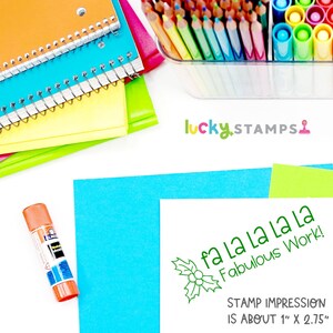 以下が含まれることがあります： 緑色の自己インク式のスタンプで、ヒイラギのデザインと「fa la la la la Fabulous Work!」というテキストが刻印されています。スタンプの印影は約2.5cm x 7cmです。
