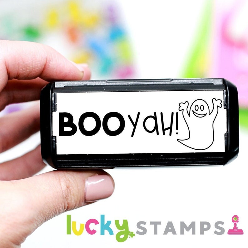 Puede incluir: Un sello autoentintado en blanco y negro con el texto "BOOyah!" y un fantasma de dibujos animados. El sello se sostiene en una mano.