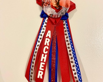 May include: Red, white, and blue ribbon with the name "ARCHER" printed on it. The ribbon is attached to a round button with a photo of a young man in a football uniform.
