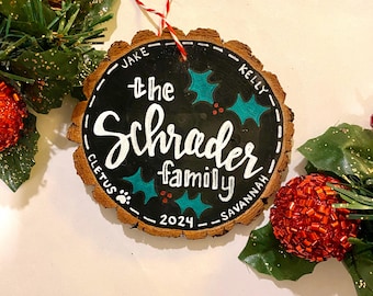 Puede incluir: Un adorno redondo de madera con una superficie de pizarra negra. El adorno está decorado con hojas de acebo verdes y bayas rojas. El texto "the Schrader family 2024" está escrito en pintura blanca en la pizarra. Los nombres "Jake", "Kelly", "Cletus" y "Savannah" están escritos alrededor del borde del adorno.