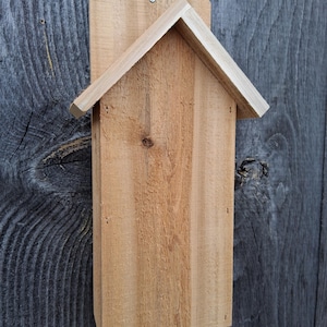 May include: A wooden bat house with a pitched roof, designed to attract bats. The house is made of natural wood and has a simple, rustic design.