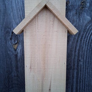 May include: A wooden birdhouse with a rectangular body and a pitched roof. The birdhouse is made of light-colored wood and is mounted on a dark wooden surface. The roof is angled, and the front has a rectangular opening.