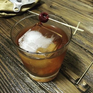 May include: A close-up of a brown cocktail in a rocks glass with a large ice cube and a maraschino cherry on a cocktail stick. The stick has a cross-shaped end.