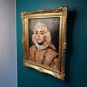 May include: A framed portrait of a woman with blonde hair wearing a crown and a white collar. The portrait is in a gold frame and is hanging on a teal wall.