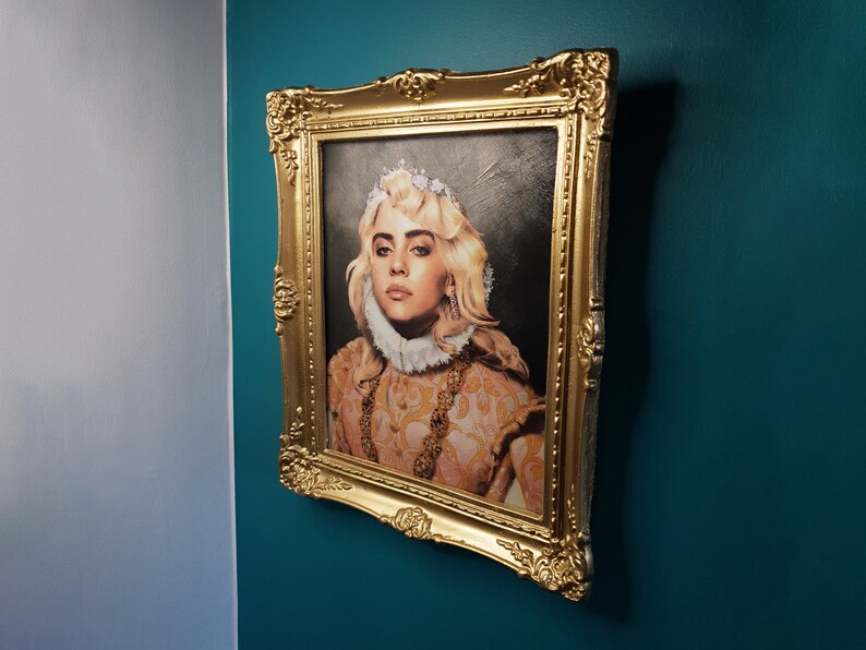 May include: A framed portrait of a woman with blonde hair wearing a crown and a white collar. The portrait is in a gold frame and is hanging on a teal wall.