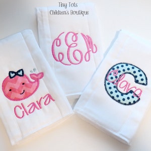 May include: Three white burp cloths with embroidered designs. One features a pink whale with a bow, another has a pink monogram, and the third has a blue and white polka dot letter C with the name Clara embroidered inside.