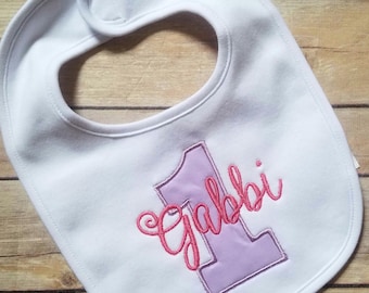 May include: White baby bib with a purple number one and the name "Gabbi" embroidered in pink.
