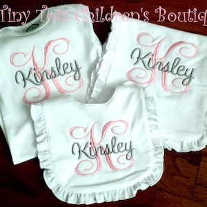 Peut inclure: Ensemble de v&ecirc;tements de b&eacute;b&eacute; blancs avec le nom "Kinsley" brod&eacute; en rose et gris. L'ensemble comprend un body, un bavoir et un bavoir d'&eacute;paule, tous avec des bords &agrave; volants. Le texte "Tiny Tots Children's Boutique" est en haut.