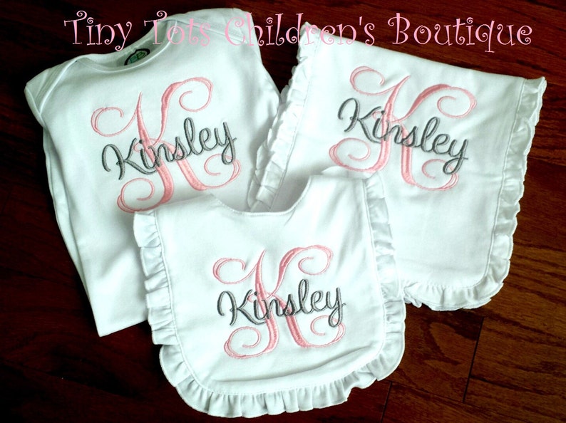 Peut inclure: Ensemble de v&ecirc;tements de b&eacute;b&eacute; blancs avec le nom "Kinsley" brod&eacute; en rose et gris. L'ensemble comprend un body, un bavoir et un bavoir d'&eacute;paule, tous avec des bords &agrave; volants. Le texte "Tiny Tots Children's Boutique" est en haut.