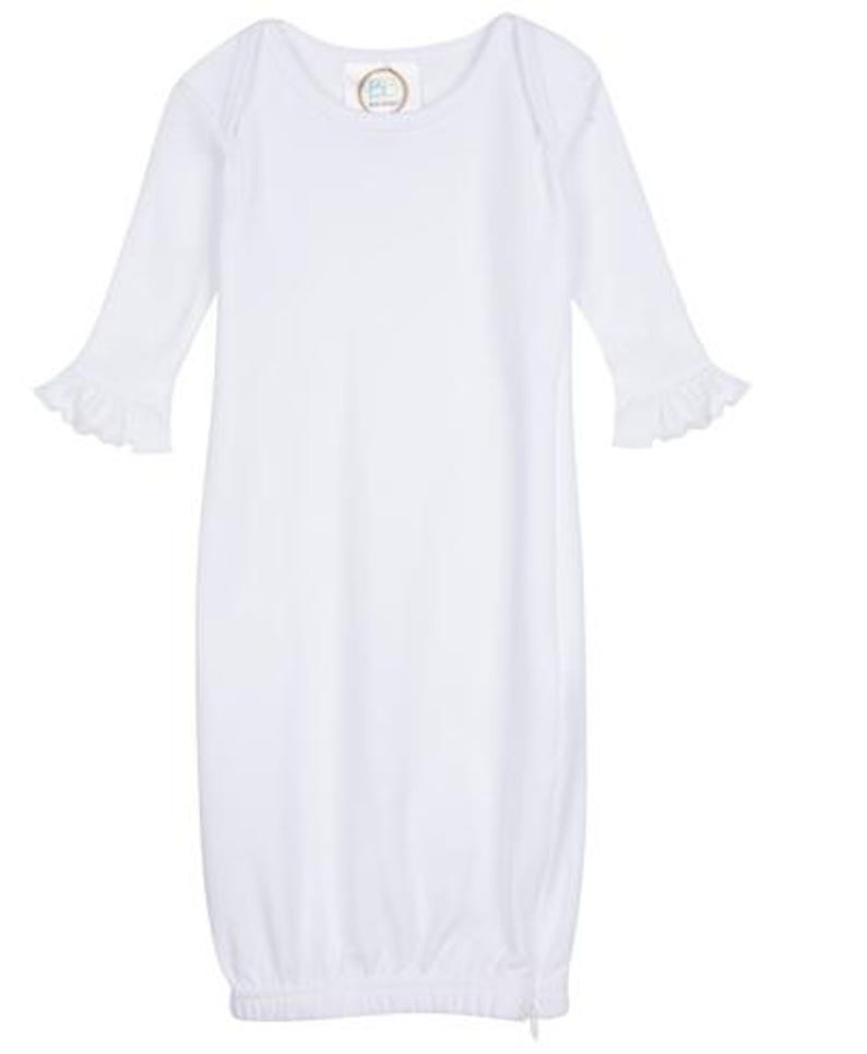 Peut inclure: Une robe de b&eacute;b&eacute; blanche avec des manches longues et des poignets &agrave; volants. La robe a un col rond et un cordon de serrage en bas. Une petite &eacute;tiquette est visible pr&egrave;s du col. La robe est en tissu doux et l&eacute;ger.