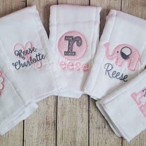 May include: Six white burp cloths with pink and gray embroidery. The designs include a monogram, a name, a letter, and an elephant.