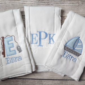 May include: Three white burp cloths with embroidered designs. The first cloth has a blue letter E with a fishing rod and fish, the second cloth has the letters EPK, and the third cloth has a blue sailboat and the name Ezra.