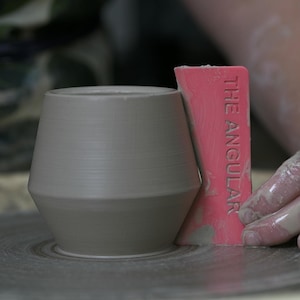 May include: A gray ceramic vase with a geometric shape is being smoothed with a pink tool that reads "THE ANGULAR". The vase is on a pottery wheel, and the artist's hand is visible.
