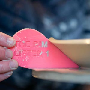 K&ouml;nnte beinhalten: Ein rosa Plastikwerkzeug mit dem aufgedruckten Text "THE RIM LIFTER 1". Das Werkzeug ist dazu gedacht, beim Anheben von Autofelgen zu helfen.