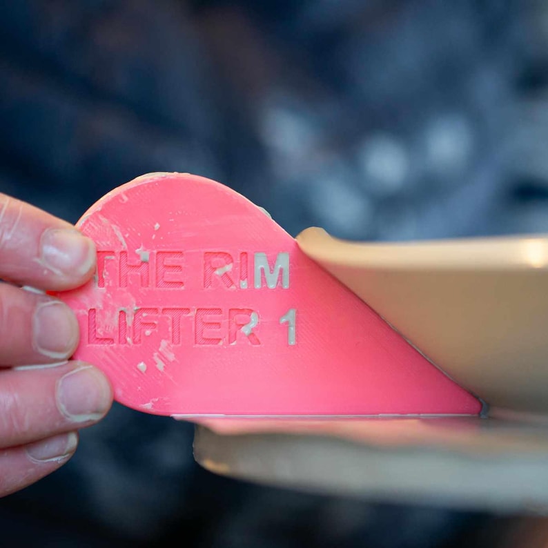 K&ouml;nnte beinhalten: Ein rosa Plastikwerkzeug mit dem aufgedruckten Text "THE RIM LIFTER 1". Das Werkzeug ist dazu gedacht, beim Anheben von Autofelgen zu helfen.