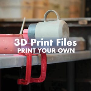 May include: A light grey ceramic mug held horizontally by a pink 3D-printed clamp attached to a red metal C-clamp. The text 3D Print Files PRINT YOUR OWN is overlaid in white. The setup is positioned on a flat surface with a pink container nearby.