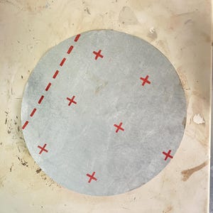 May include: A circular, light gray object with red cross marks and a dashed red line on the left side. The object is set against a light beige background, suggesting a craft or design project.