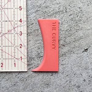 May include: A pink plastic tool shaped like a curved line with the text "THE CURVY" printed on it. The tool is next to a ruler.