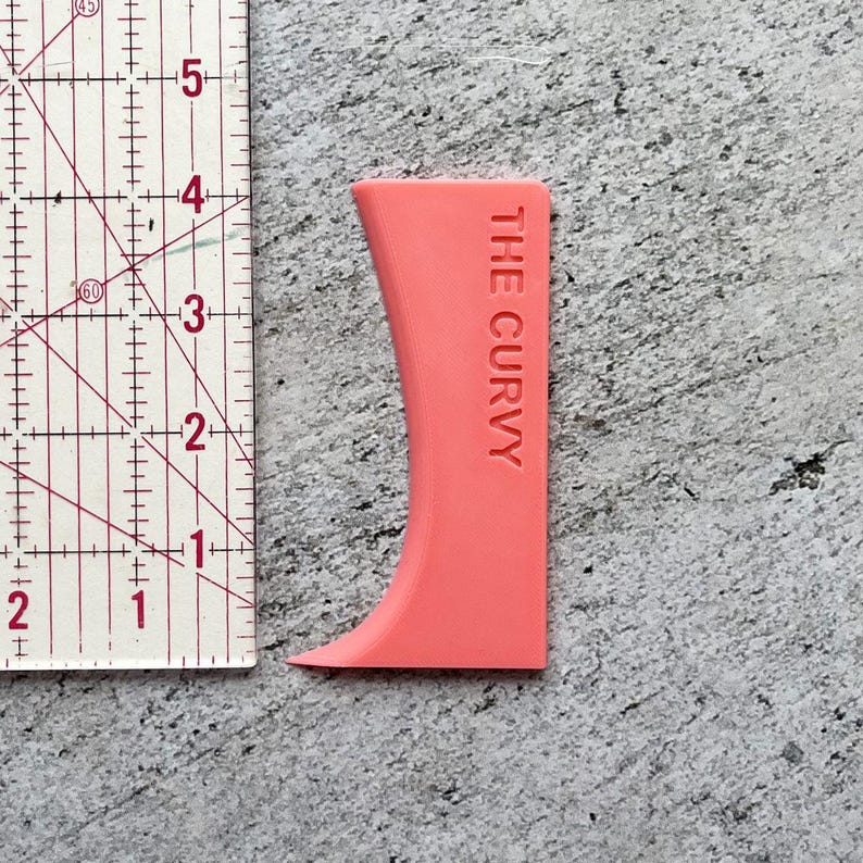 May include: A pink plastic tool shaped like a curved line with the text "THE CURVY" printed on it. The tool is next to a ruler.