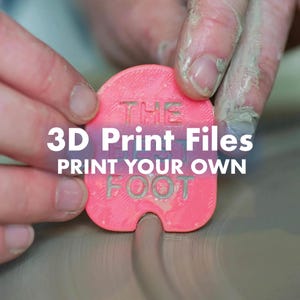 Puede incluir: Una herramienta de alfarería rosa brillante, redondeada e impresa en 3D con una superficie texturizada. La herramienta presenta el texto en relieve THE BEST FOOT. Se sostiene contra una superficie de arcilla gris con una muesca curva.