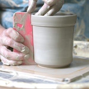 May include: A light grey ceramic cylinder is being shaped on a pottery wheel. A pink rib tool is used to smooth the exterior. The potter's hands and tool are covered in wet clay, showing the pottery process.