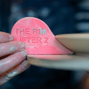 K&ouml;nnte beinhalten: Ein rosa Keramikwerkzeug mit der Aufschrift "THE RIM LIFTER 2", das zum Anheben des Randes von Keramik verwendet wird.