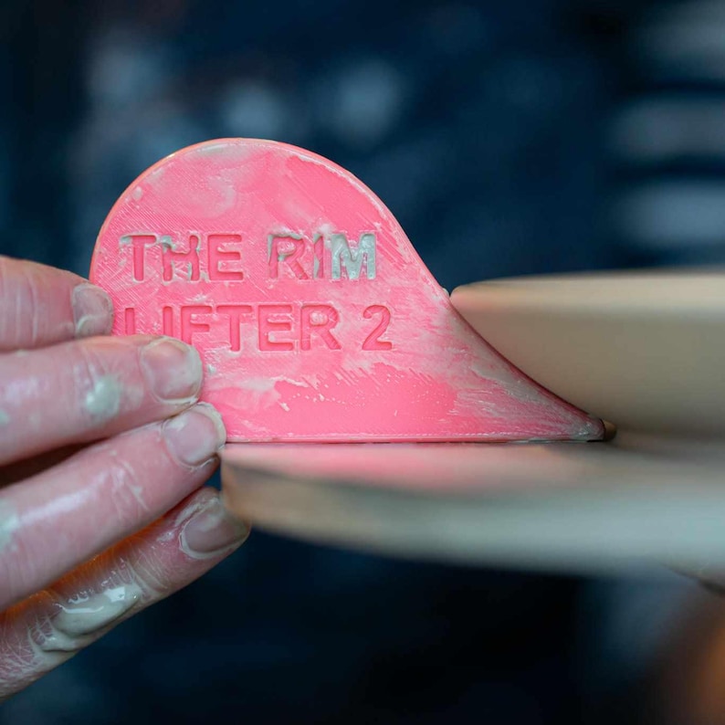 K&ouml;nnte beinhalten: Ein rosa Keramikwerkzeug mit der Aufschrift "THE RIM LIFTER 2", das zum Anheben des Randes von Keramik verwendet wird.