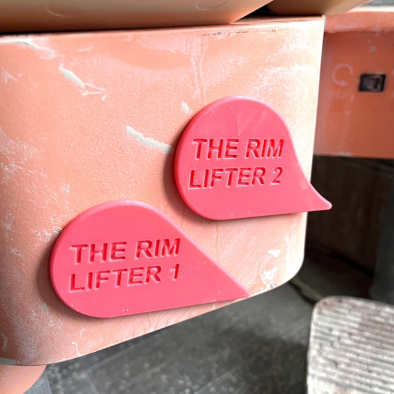 K&ouml;nnte beinhalten: Zwei rote Plastiklaschen mit dem Text "The Rim Lifter 1" und "The Rim Lifter 2" auf einer hellorangen Oberfl&auml;che.