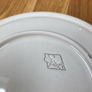 May include: White ceramic plate with a raised, diamond-shaped logo that reads "Spade" in a cursive font.