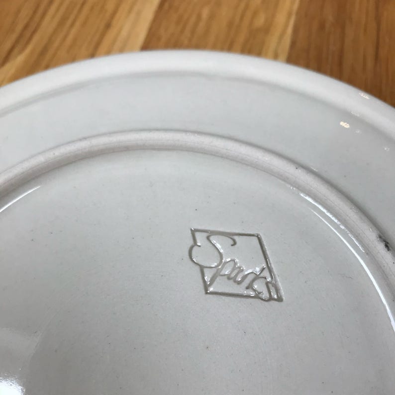 May include: White ceramic plate with a raised, diamond-shaped logo that reads "Spade" in a cursive font.