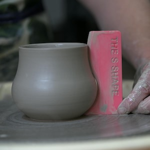 May include: A gray ceramic vase with a rounded body and a wide opening. A pink tool with the text "THE S SHAPE" is positioned next to the vase. The vase is on a pottery wheel.
