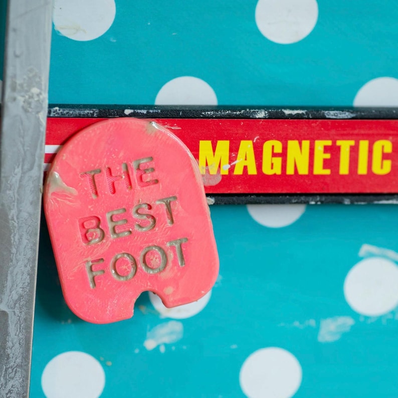 May include: A red plastic magnet with the words "The Best Foot" printed on it. The magnet is attached to a black and red magnetic strip with the word "Magnetic" printed on it.