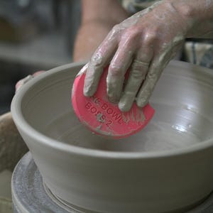 May include: A potter's hands shaping a grey ceramic bowl. A pink tool, marked "THE BOWL BOSS 2", is used. The potter's hands are covered in wet clay. The bowl sits on a pottery wheel, ready for the next stage.